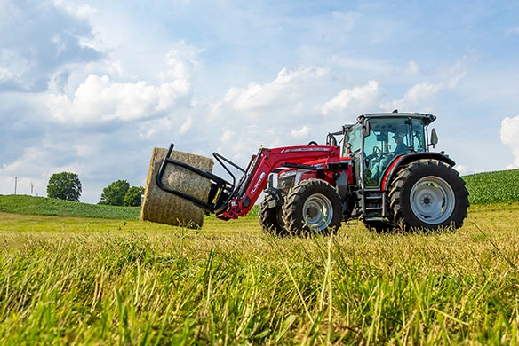 Massey Ferguson 5M tractor with front loader attachment carrying a large round hay bale in a field.