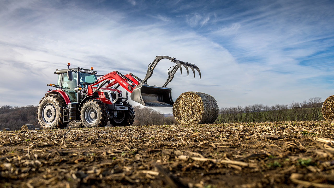 Massey Ferguson 6S Series tractor lifting a round bale in a harvested field under a partly cloudy sky.