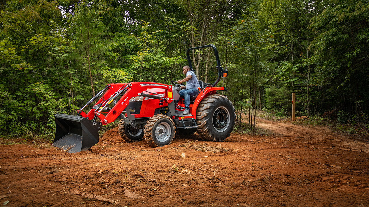 Man operating a Massey Ferguson 1800M compact tractor with front loader in wooded area, ideal for land clearing and trail work.