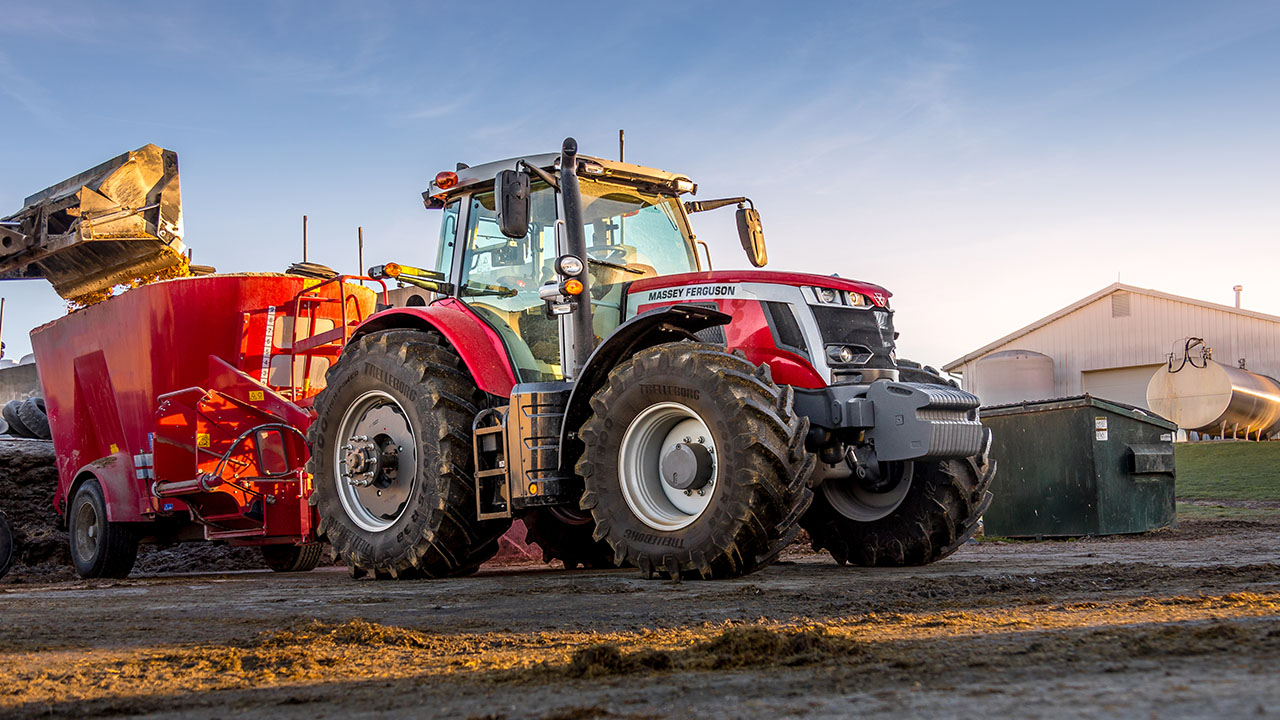 Massey Ferguson 7S Series tractor hauling a red feed mixer on a livestock farm at sunrise.
