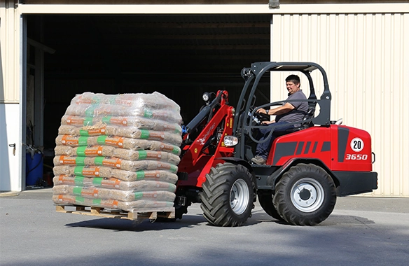 Thumbnail for Schäffer 3650 loader carrying a full pallet of bagged materials into a warehouse with a pallet fork attachment.