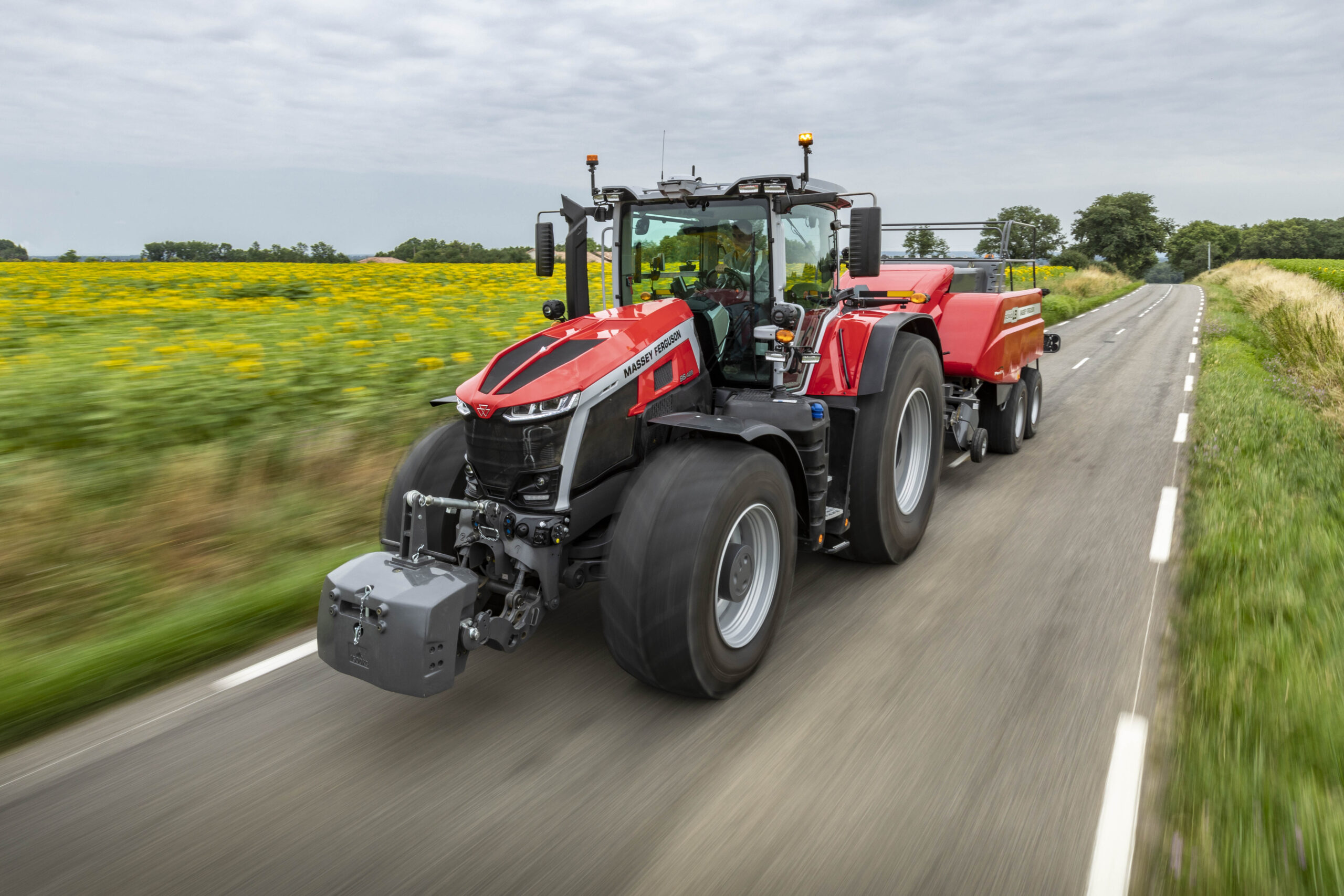 Massey Ferguson 9S Series tractor towing a square baler on a rural road next to sunflower fields under a cloudy sky.
