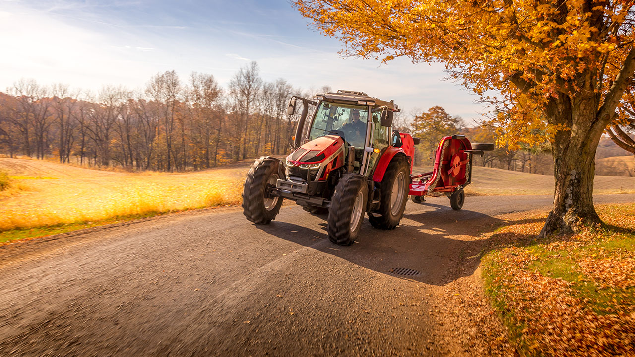 Massey Ferguson 5S Series tractor hauling a rotary cutter on a scenic country road in autumn, ideal for hay and land maintenance.