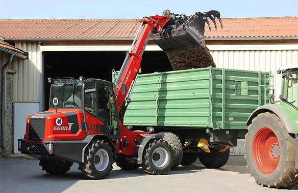 Schäffer 6680 T telescopic loader with grapple bucket loading organic material into a green farm trailer.