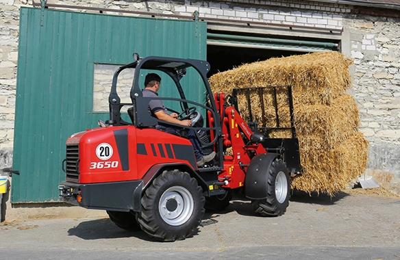 Schäffer 3650 compact loader stacking rectangular straw bales outside a barn using a bale fork.