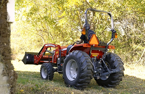 Rear angle of Massey Ferguson 2E Series compact tractor equipped with front loader and rear three-point hitch, positioned in a wooded rural setting, emphasizing versatility for small farms and property maintenance.