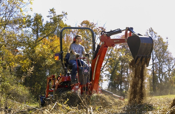 Massey Ferguson 1GC Series compact tractor with backhoe digging in grassy, wooded area.