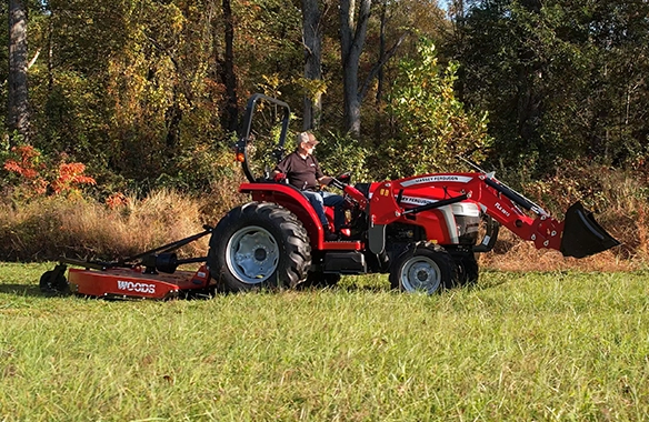Operator driving a Massey Ferguson 2E Series compact tractor with front loader and rear rotary cutter attachment across a pasture, demonstrating mowing performance and all-day operator comfort.