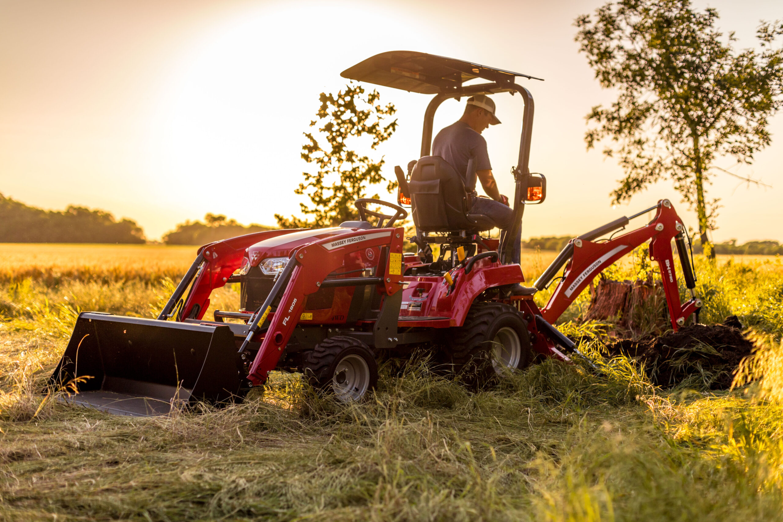 A red Massey Ferguson GC1700 Series sub-compact tractor with a backhoe attachment digging out a stump in a grass field.
