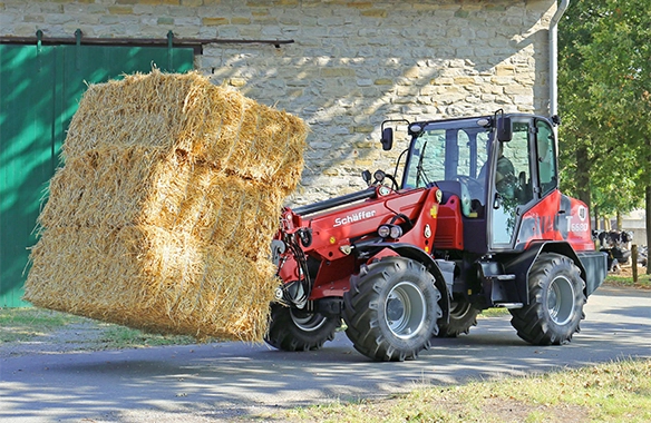 Schäffer 6680 T telescopic wheel loader transporting stacked straw bales outside a stone barn using a bale fork.