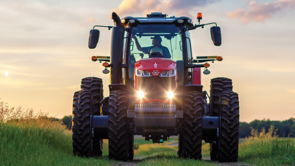Massey Ferguson 8700S Series tractor with duals driving on a grassy farm path at sunset, headlights on for early evening fieldwork.