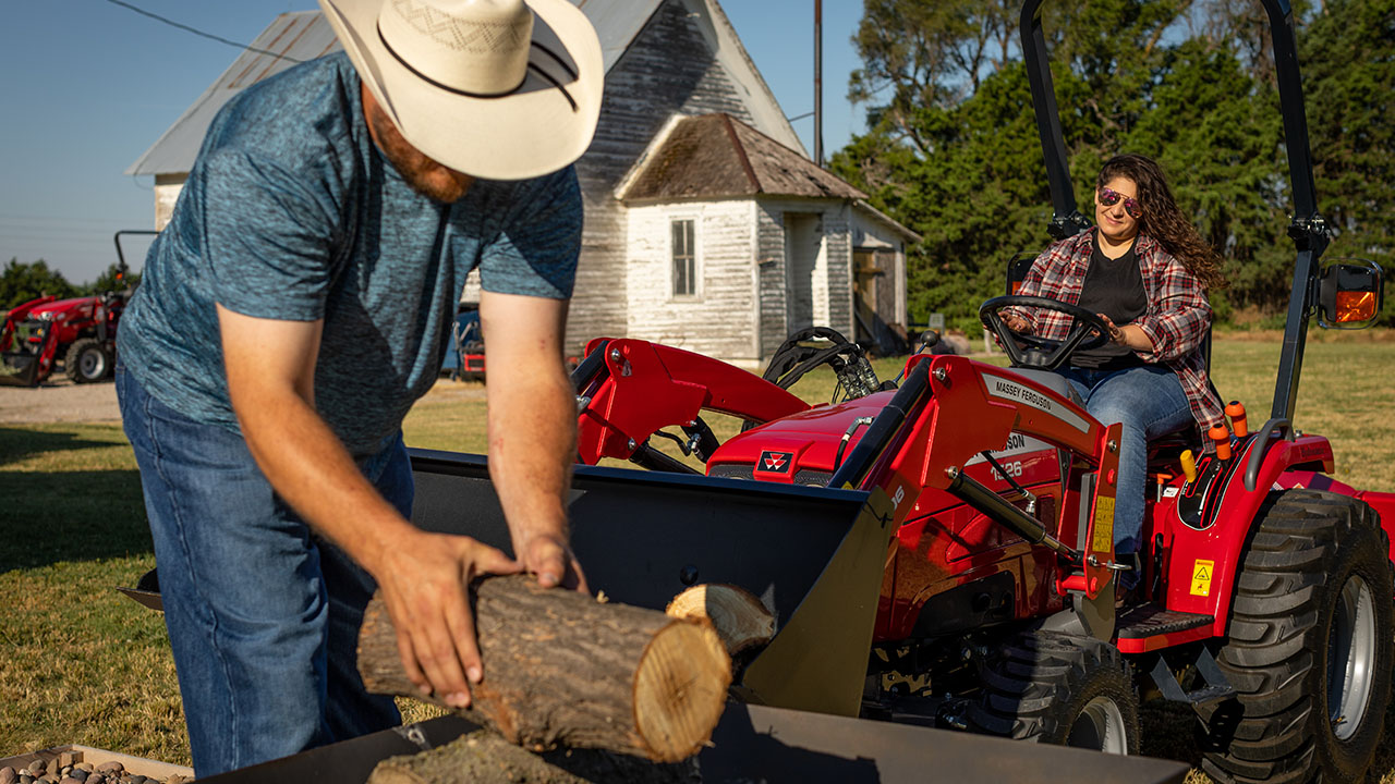 Woman driving Massey Ferguson 1526 tractor while man loads firewood into front loader on rural property.