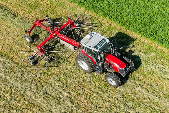 Massey Ferguson 5M utility tractor raking hay with a rotary rake attachment in a field.