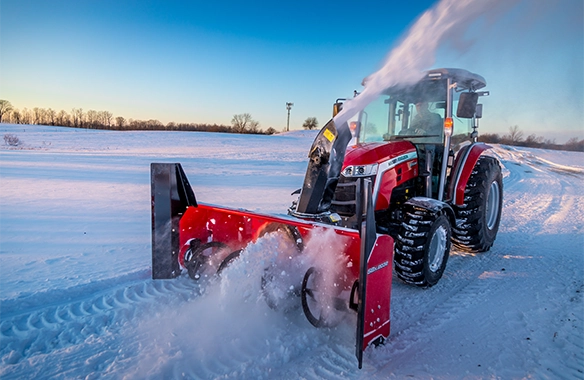 Front view of Massey Ferguson 2M Series compact tractor equipped with snow blower, actively removing snow from a driveway in winter conditions at sunrise, highlighting cold-weather performance.