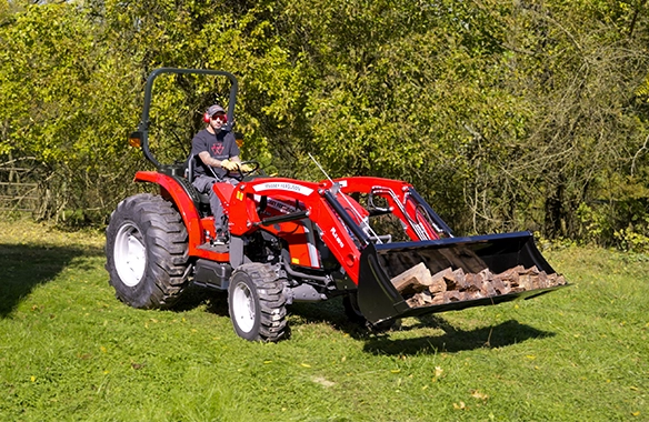 Man operating a Massey Ferguson 2E Series compact tractor with front loader carrying firewood, ideal for rural landowners managing woodlots, chores, and seasonal tasks with heavy loads.