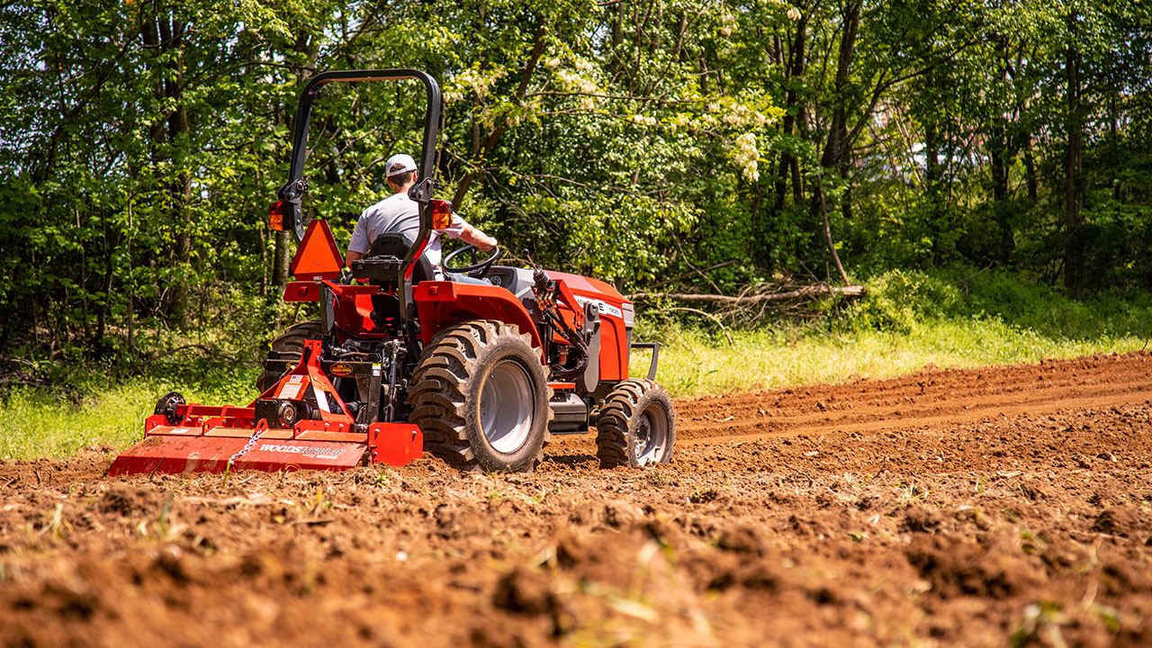 Thumbnail for Massey Ferguson 1800E tractor with Woods rotary tiller cultivating a field, operated by man in cap near wooded area.