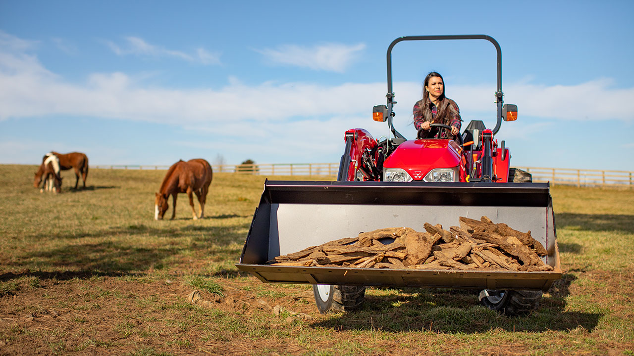 Woman operating Massey Ferguson 2800E Series compact tractor with loader carrying wood in horse pasture, ideal for farm chores.