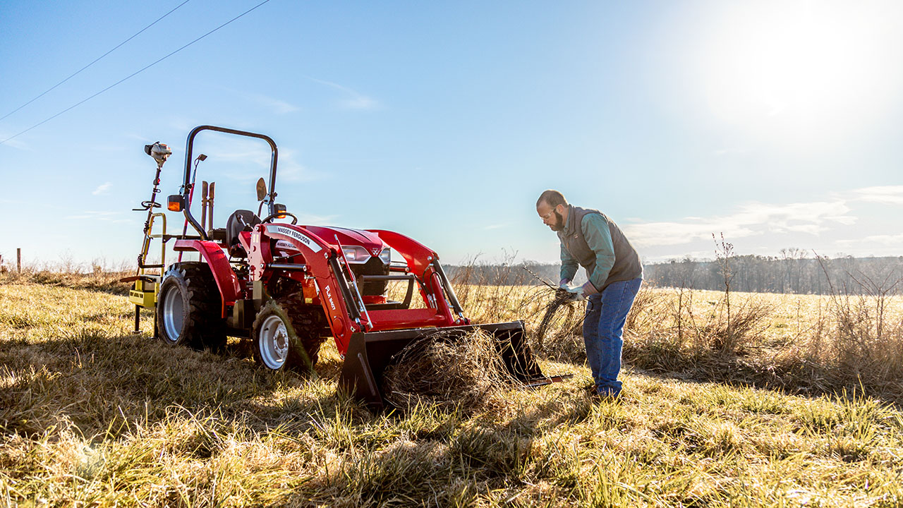 Man clearing brush beside a Massey Ferguson 1800 Series compact tractor on open farmland under sunny sky.