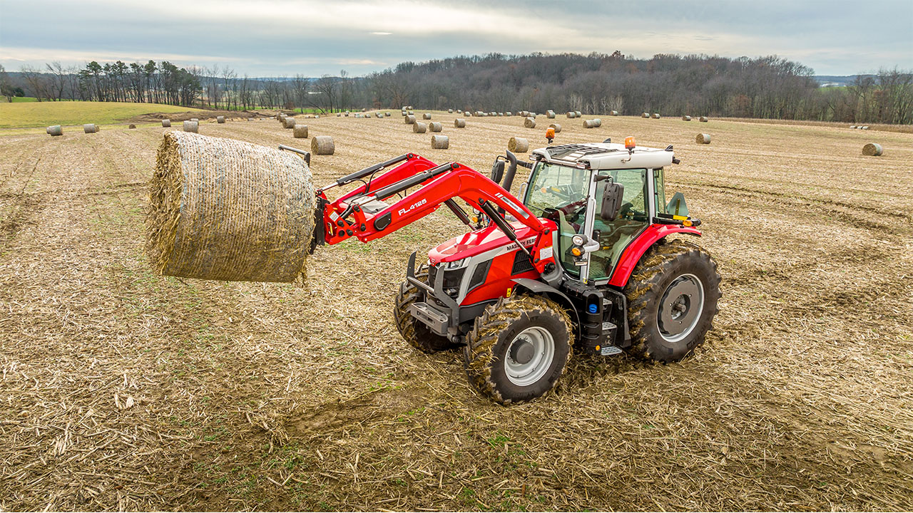 Massey Ferguson 6S Series tractor lifting a round bale in a harvested field under a partly cloudy sky.