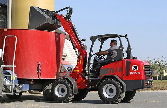 Schäffer 3650 loader filling a red feed mixer wagon with silage on a farm.