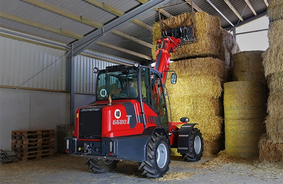 Schäffer 6680 T loader stacking round hay bales inside a barn with extended telescopic arm and bale grab.