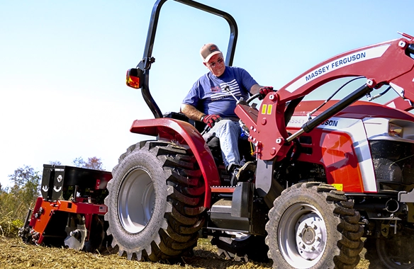 Man operating a Massey Ferguson 1E Series compact tractor with a front loader and rear rotary tiller attachment, showcasing its performance and versatility in field preparation tasks.